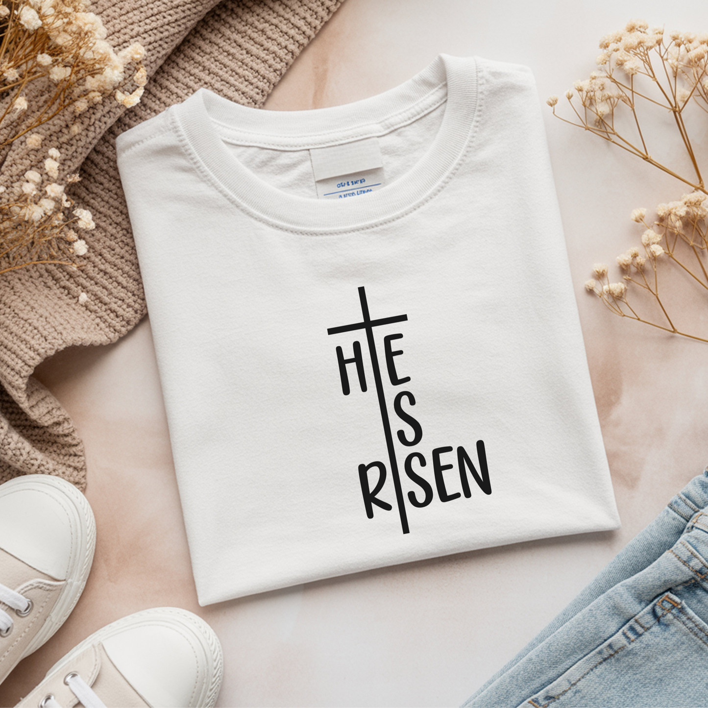 White t-shirt with black 'He is Risen' cross design, laid flat with neutral decor