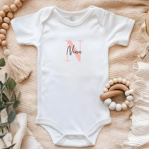 Bügelbild Baby für Body Mädchen, personalisiert mit Großbuchstaben in Rosa und Name in schwarzer Schrift. Das Design hat eine Breite von 7 cm und eignet sich perfekt für den kleinen Body. Ein tolles individuelles Geschenk für das Baby