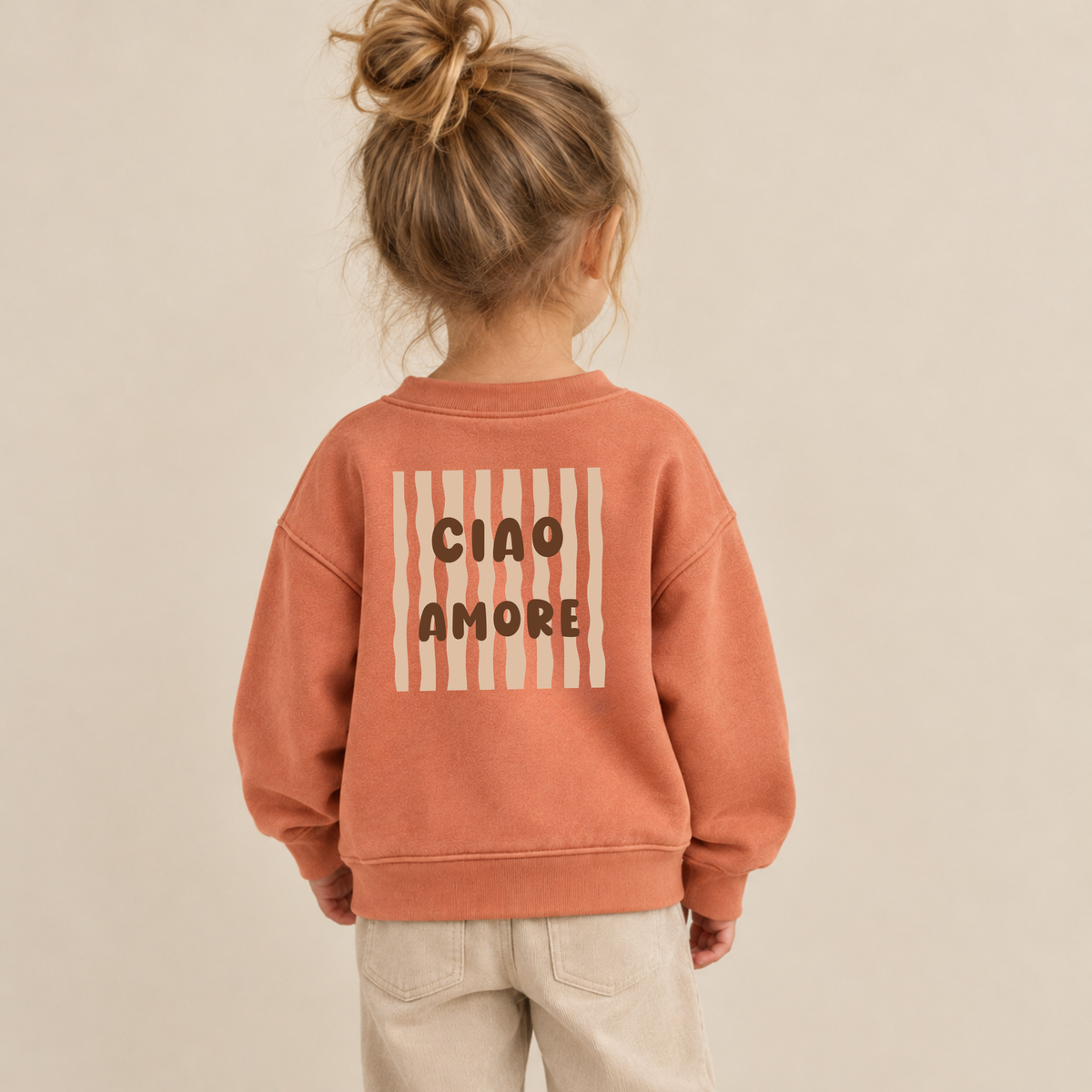 Bügelbild „Ciao Amore“ mit braunem Schriftzug und beigen Wellenstreifen, aufgebügelt auf Rückseite von terracotta Kinderpullover. Bügelmotiv für Rücken oder Front