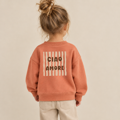 Bügelbild „Ciao Amore“ mit braunem Schriftzug und beigen Wellenstreifen, aufgebügelt auf Rückseite von terracotta Kinderpullover. Bügelmotiv für Rücken oder Front