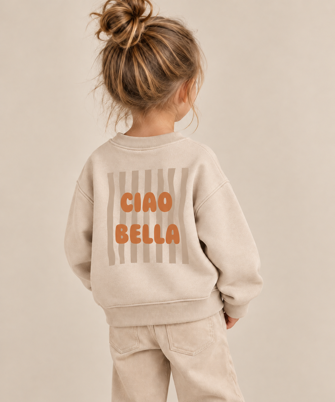 Bügelbild „Ciao Bella“ mit orangem Schriftzug und beigen Wellenstreifen, aufgebügelt auf Rückseite von beigem Kinderpullover. Bügelmotiv für Mädchen und Kinder.